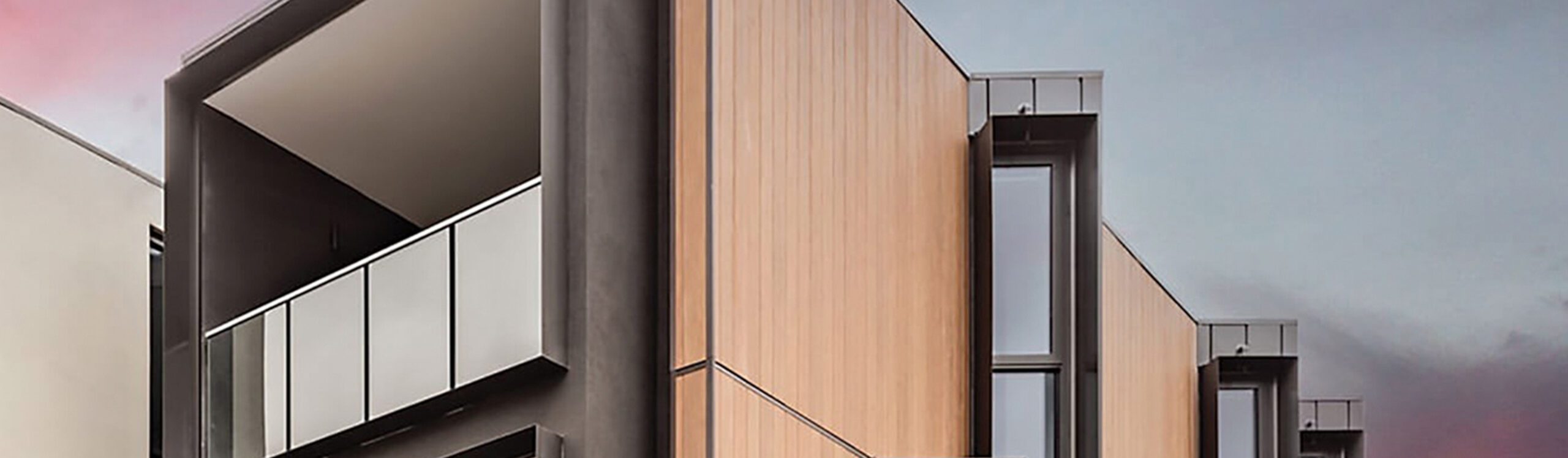 Modern architectural building with a minimalist design featuring large windows, wooden paneling, and grey metallic accents. The structure includes a balcony with a glass railing, set against a sky transitioning from pink to blue.