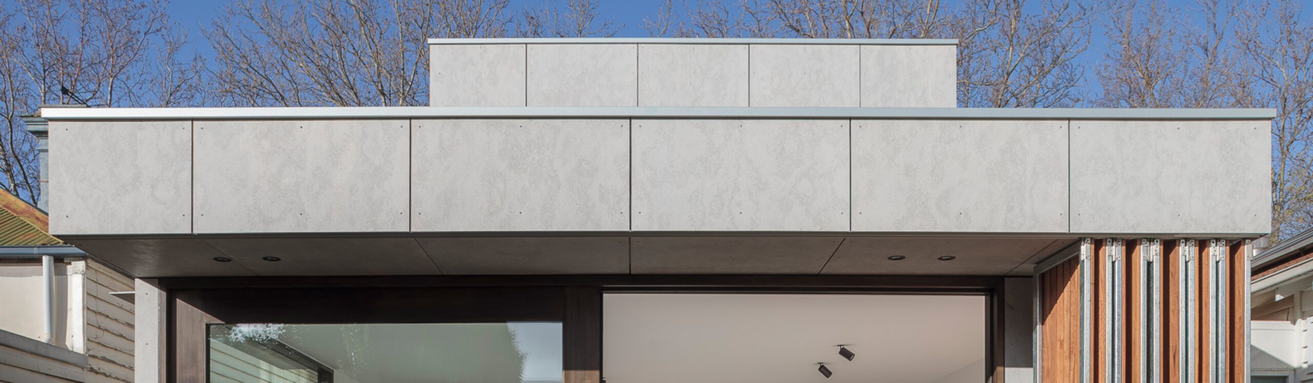 A modern building facade with a flat roof and light grey paneling. The structure features large glass doors and windows, and a clear blue sky is visible in the background. Sparse trees without leaves stand behind the building.