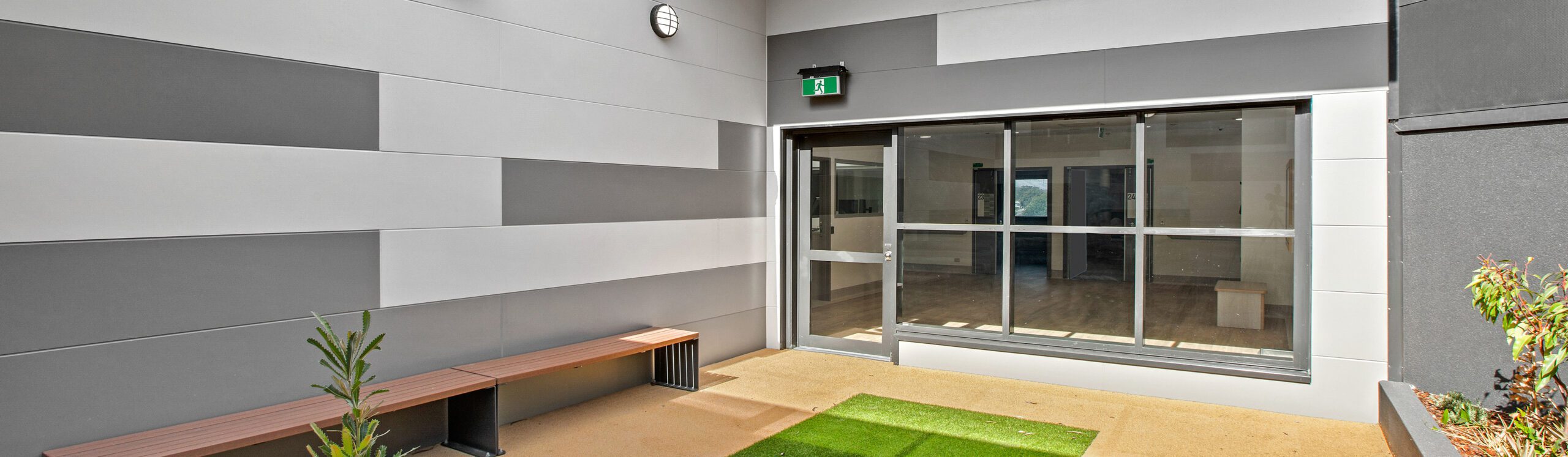 A modern indoor courtyard with geometric wall panels in shades of gray and white. The area features a wooden bench, a small green artificial grass patch, potted plants, and a large glass sliding door leading into a room. An emergency exit sign is visible above the door.