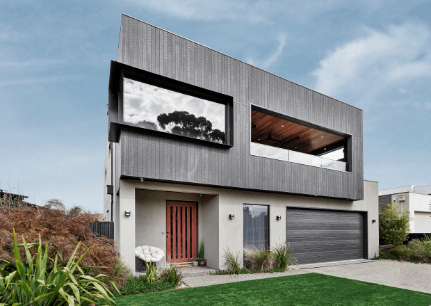 A modern two-story house with a mix of dark and light exterior finishes. Large windows on both floors, a wooden front door, and a gray garage door. The front yard features green grass, minimal landscaping, and a concrete pathway leading to the entrance.