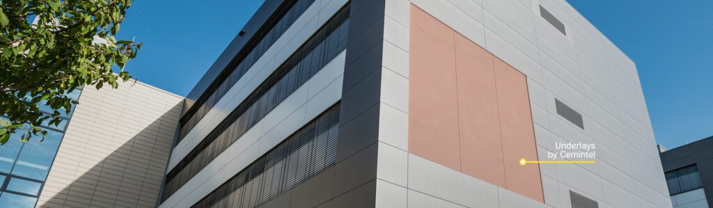 A modern multi-story building with a light grey and beige exterior stands against a blue sky. A small portion of the facade is labeled "Underlays by Cemintel." A leafy tree is partially visible in the foreground to the left.