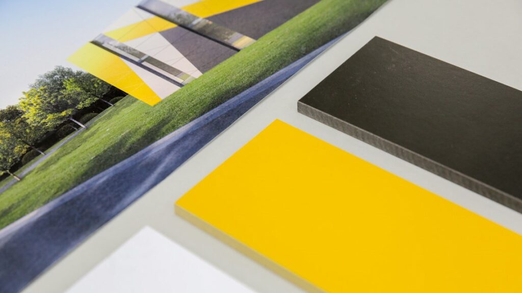 Close-up of a color swatch of panels laying on a table, including yellow, black, and white samples. In the background, there is an image of a modern building with similar colors, surrounded by green grass and trees.