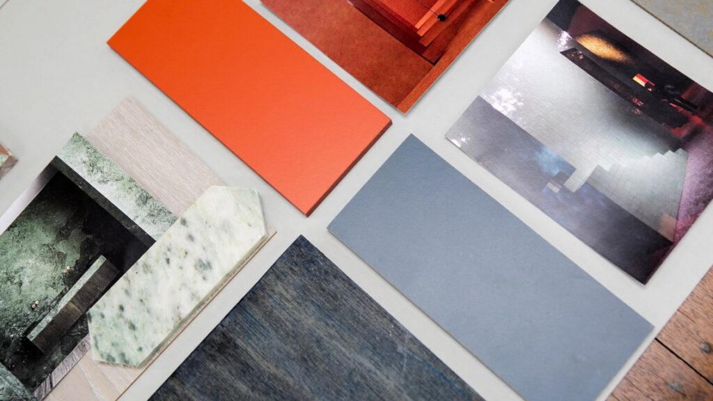 A layout of various material and color samples is spread on a flat surface. The samples include orange, gray, and textured tiles, alongside images of interior spaces, wood finishes, and a stone-textured piece. The arrangement showcases different design options.