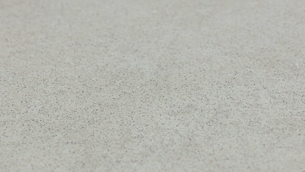 A close-up view of a textured surface, featuring a beige background with numerous tiny black and brown specks scattered uniformly across it. The specks create a subtle, granulated appearance on the smooth surface.