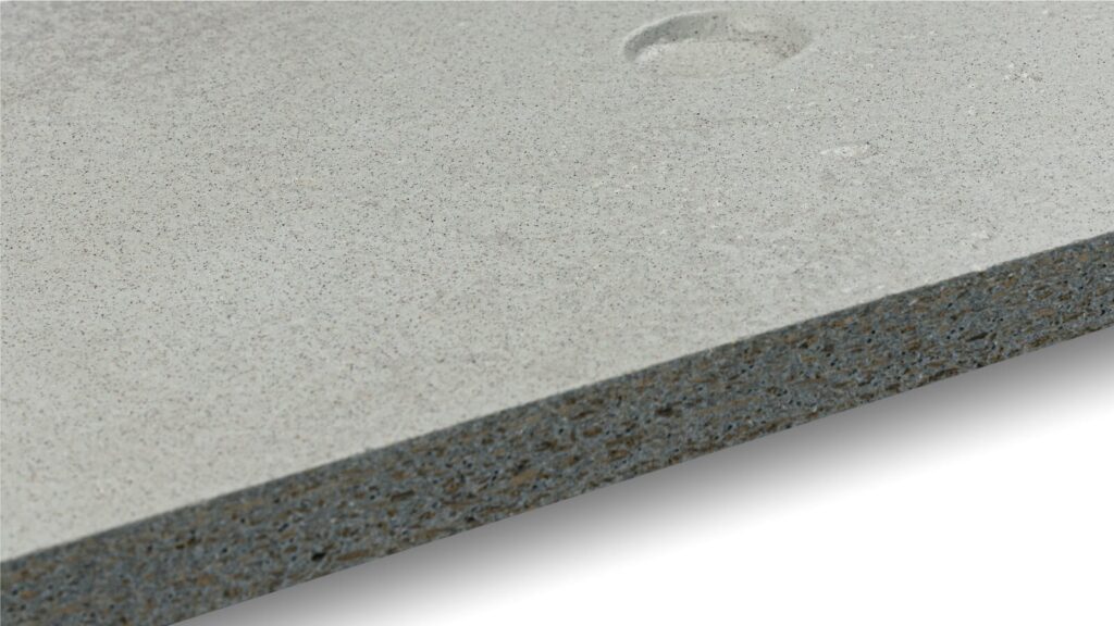 Close-up image of a textured concrete surface. The concrete is gray with small specks throughout, displaying a rugged, unfinished appearance. There is a circular indentation on the top right, adding to the raw, industrial feel of the material.