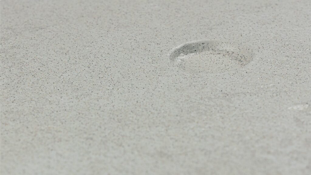 A close-up of a light grey sandy surface with a single shallow indentation near the center. The texture of the sand is smooth with scattered tiny specks, creating a subtle, grainy appearance.