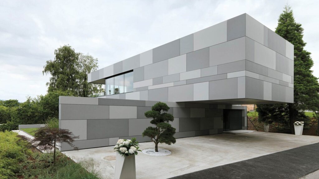 Modern two-story house with a sleek, geometric design and gray panel exterior. An overhanging second floor creates a covered driveway. The yard features a small tree, shrubs, and a flower arrangement near the entrance. Lush greenery surrounds the property.