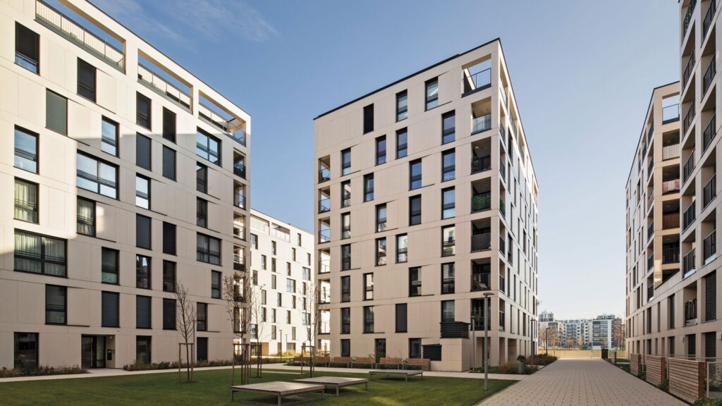 A view of modern apartment buildings with multiple floors and large windows. The buildings are arranged around a courtyard with green grass, benches, and young trees. The sky is clear and blue, and the pathway through the courtyard is lined with street lamps.