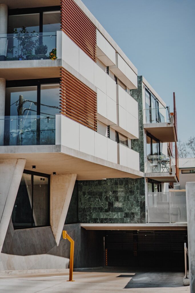 Modern multi-level apartment building with a sleek, minimalist design. The structure features large windows, balconies with glass railings, and a mix of concrete, wood, and green marble facade elements. There's an entrance to an underground garage visible at the bottom.