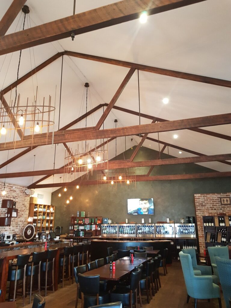 A cozy restaurant with wooden beams and hanging lights on the ceiling. Tables and chairs are arranged neatly, with a wine display and a large TV on one wall. The floor is wooden, and some brick accents add to the rustic charm.