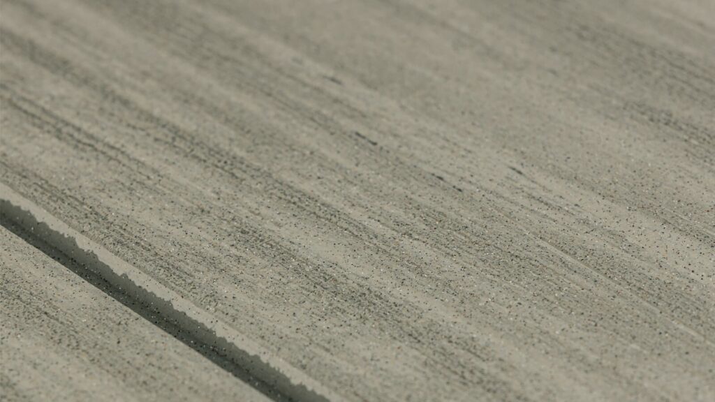 Close-up of a concrete surface with textured, slightly uneven lines running horizontally. The color is a mix of light gray tones with small, scattered specks creating a granular appearance, adding a subtle variation to the surface.