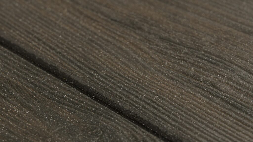 Close-up view of a textured surface resembling dark wood grain. The surface displays linear grooves and subtle patterns, giving it a natural and rustic appearance. The color is a deep brown, enhancing the wood-like impression.