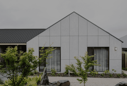 Modern house with a gabled roof and large windows, surrounded by landscaped greenery and a gravel path. The exterior walls are light gray, contrasting with the darker roof. Overcast sky in the background.